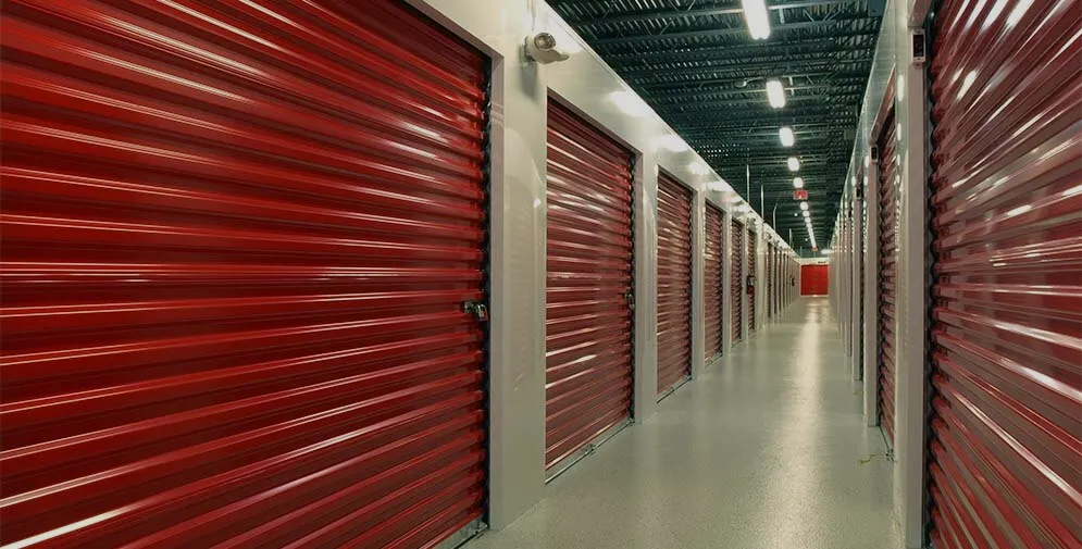 A long corridor flanked by multiple red storage units with roll-up doors. The facility appears modern and clean, with a polished concrete floor that reflects the fluorescent lights overhead.